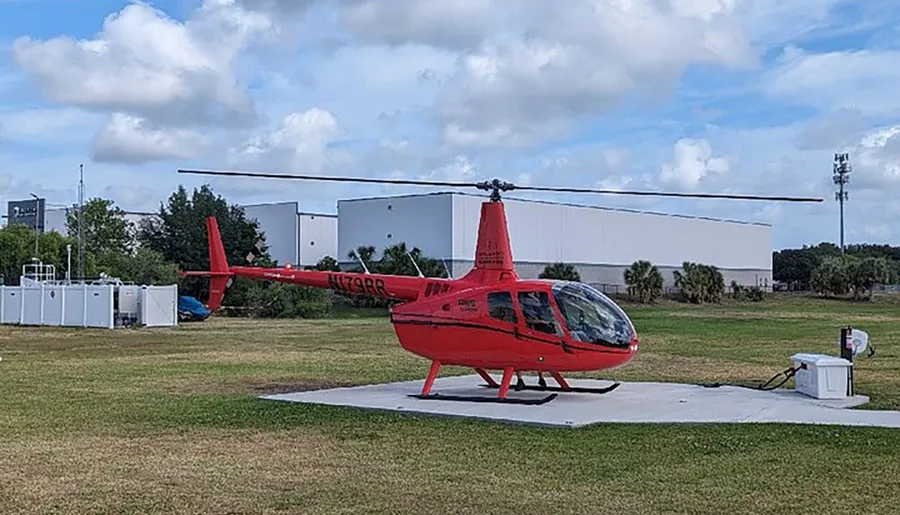 A red helicopter is parked on a landing pad in an open field with industrial buildings in the background.