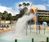 The image shows a vibrant outdoor water park with a playful childrens splash zone surrounding sun loungers fake rock formations and a scenic backdrop of trees and a lake