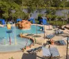 The image shows a vibrant outdoor water park with a playful childrens splash zone surrounding sun loungers fake rock formations and a scenic backdrop of trees and a lake