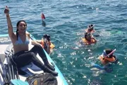 A person is cheerfully raising her arm on a boat while others with snorkeling gear swim in the sea nearby.