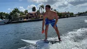 A person is wake surfing on a wave created by a boat on a sunny day with palm trees and houses in the background.