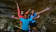 Three joyful individuals are posing with arms raised in a cave-like setting, expressing happiness or excitement.