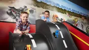 Two children are joyfully playing with toy cars on a specially designed race track that emulates a real racing scenario, with a dynamic racing-themed mural in the background.