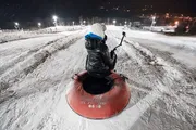 A person is sitting atop a red snow tube at night, ready to slide down a snowy hill, while holding a camera or smartphone on a stick to capture the moment.