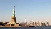The Statue of Liberty stands prominently in the foreground with the Manhattan skyline, including One World Trade Center, in the background under a clear sky.