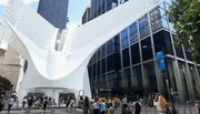 The image shows a bustling urban scene with people walking in front of the distinctive, modern, white ribbed structure of the Oculus in New York City.