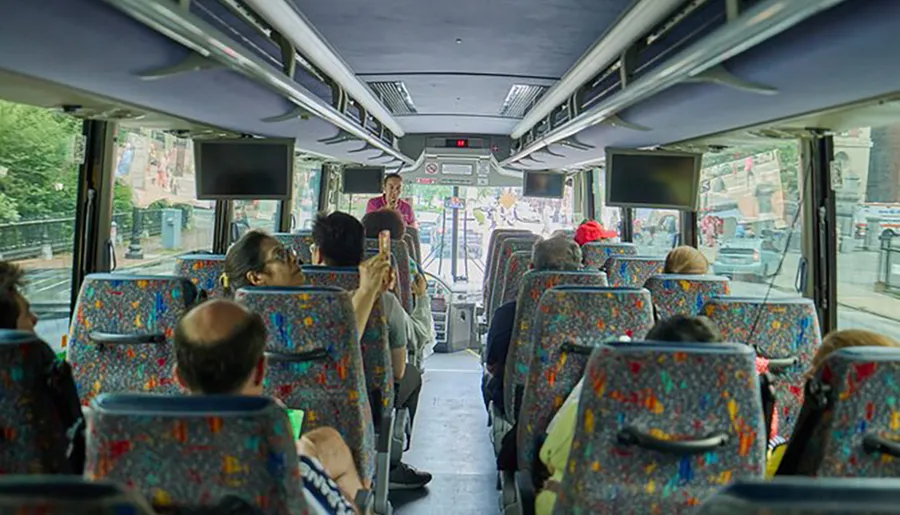 Passengers are seated on a colorful bus with a tour guide standing in the aisle presumably giving information or instructions.