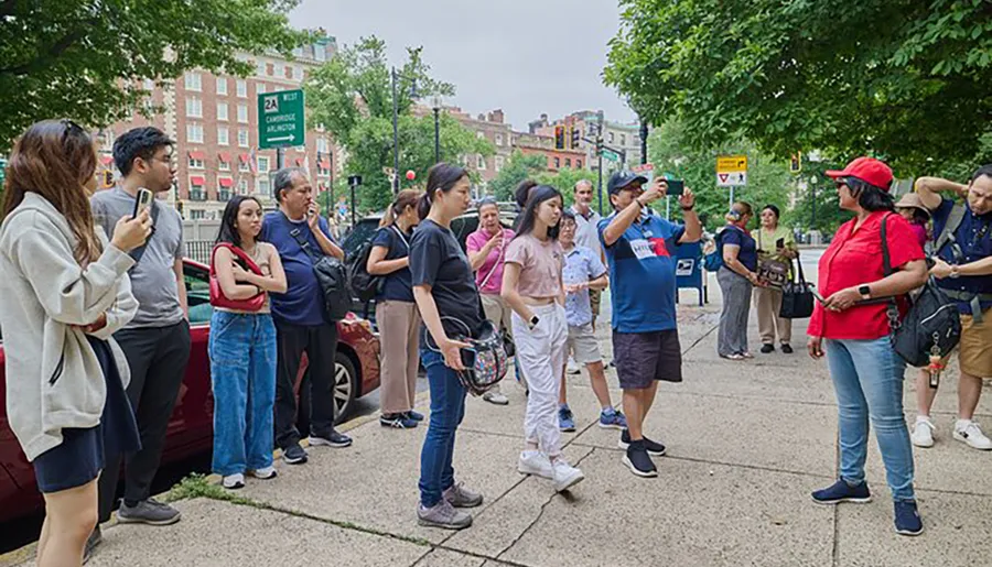 A group of attentive people is listening to a person gesturing and explaining something, possibly a tour guide, on a city street corner.