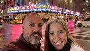 A smiling man and woman are taking a selfie in front of the brightly lit marquee of Radio City Music Hall at night.