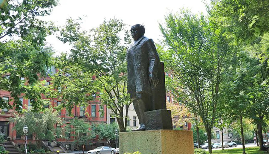 The image shows a statue of a historical figure standing on a pedestal in an urban park with lush greenery and redbrick buildings in the background.