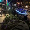 The image shows a vibrant evening scene with people riding in a boat under illuminated trees alongside a riverwalk, with others strolling and enjoying the atmosphere.