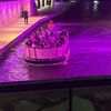 A group of people enjoy a boat tour under a vividly lit purple underpass on a riverwalk at night.