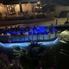 A boat adorned with blue lights carries passengers along a river at night, with spectators on the riverside walkway.