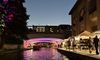 The image captures an evening scene of a vibrant urban riverwalk with pedestrians, market stalls, and an illuminated bridge reflecting colored lights onto the water.