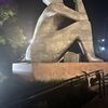 The image shows a large, abstract metal sculpture illuminated at night, possibly situated in a public space with vegetation around it.