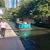 A boat full of passengers cruises along a river beside a walkway where a person is walking and kiosks display souvenirs.