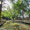 The image shows a serene urban river walk flanked by lush green trees, with sunlight filtering through the foliage.