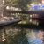 The image shows a tranquil urban riverwalk scene with dappled sunlight filtering through trees, reflecting off the water and a bridge in the background, with some people visible walking alongside the river.