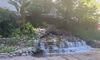 The image depicts a serene outdoor pond with a small waterfall surrounded by lush vegetation and a building in the background.