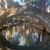 An arched stone bridge spans a reflective waterway lined with trees and buildings under a clear sky.