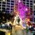 A stone monument stands in front of a tall building, with a tree covered in colorful Christmas lights nearby.