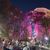 A group of people admire colorful lights decorating the trees along a riverside at night.