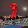 The image features a large, illuminated red sculpture against a night sky with people standing on a stone bridge, decorated with a Christmas wreath and lit up in colorful lights.