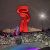 A large red, spiraling sculpture is illuminated at night above a stone bridge decorated with holiday lights and a wreath, with people gathered nearby.