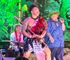 A performer in traditional attire is energetically dancing with a stick, while a person beside him appears to be singing or speaking into a microphone, set against a vibrant, tropical-themed backdrop.