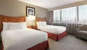 This is a neatly arranged hotel room with two double beds, a framed picture hanging above a nightstand, and a large window with sheer curtains.