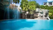 The image shows a serene swimming pool with a cascading artificial waterfall, surrounded by tropical vegetation and architectural structures, conveying a resort-like atmosphere.
