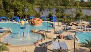 The image shows a vibrant outdoor water park with a playful children's splash zone, surrounding sun loungers, fake rock formations, and a scenic backdrop of trees and a lake.