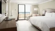 The image shows a tidy and modern hotel room with a large bed, minimalist furniture, and a balcony overlooking the ocean.