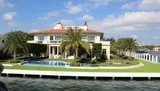 The image showcases an elegant, two-story waterfront mansion with a swimming pool, palm trees, and a well-manicured lawn, viewed from across the water.