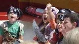 A group of children, dressed as pirates and sporting eye patches and bandanas, are joyfully engaging in a themed activity, with one girl triumphantly raising her fist and holding a toy sword.