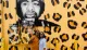 A couple is posing for a photo in front of a vibrant yellow wall featuring an artistic mural with black and orange leopard print and a large portrait.