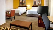 The image shows a modern hotel room with two beds, a work desk with a chair, a television, and warm, inviting decor.