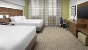 The image shows a modern hotel room with two beds, a work desk with a chair, a flat-screen TV, and a patterned curtain covering a window.