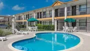 This image shows a sunny outdoor scene with a clear blue swimming pool in the foreground and a two-story motel with balcony access rooms in the background, complete with umbrellas and seating arrangements around the pool area.