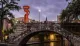 An old stone bridge spans a river in an urban setting, illuminated by the glow of streetlights and an orange sculpture at dusk.