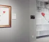 The image features an interior wall of a gallery with opposite displays of an artwork depicting a girl reaching for a heart-shaped balloon one is a framed print and the other is a larger mural-style representation