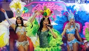The image shows performers in vibrant, elaborate costumes, with the central figure singing or speaking into a microphone on a stage set with tropical-themed decorations.