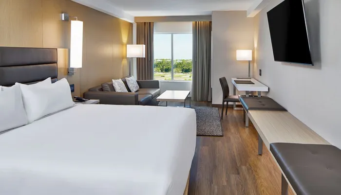 This image shows a modern hotel room with a large bed a seating area a desk and a flat-screen TV featuring a neutral color palette and a view of the outside through large windows