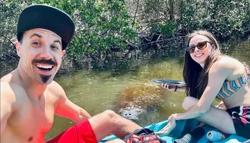 A smiling man and woman are enjoying kayaking together, with a manatee visible in the water near them.