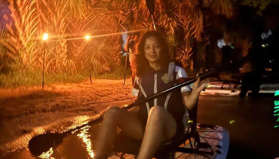 A person is sitting on a paddleboard on the water at night, illuminated by nearby warm lights.