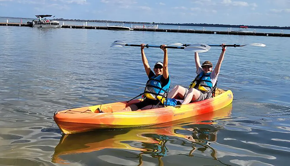 Two people are joyfully raising their paddles while seated in a tandem kayak on a calm body of water