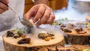 A chef is meticulously plating a dish with tweezers amid a smoky presentation on wooden surfaces, suggestive of an upscale gourmet experience.