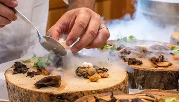 A chef is meticulously plating a dish with tweezers amid a smoky presentation on wooden surfaces, suggestive of an upscale gourmet experience.
