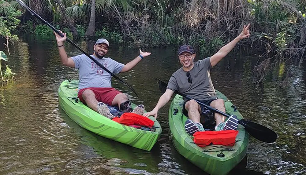 Two people are joyfully kayaking in a calm waterway surrounded by lush greenery