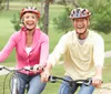 Two smiling people wearing cycling helmets are riding bicycles together in a park-like setting
